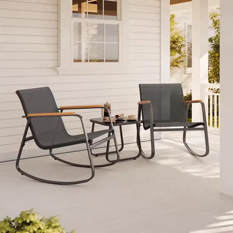 Two black mesh rocking chairs with wooden armrests and a small glass-top table sit on a white porch with greenery and a window in the background.