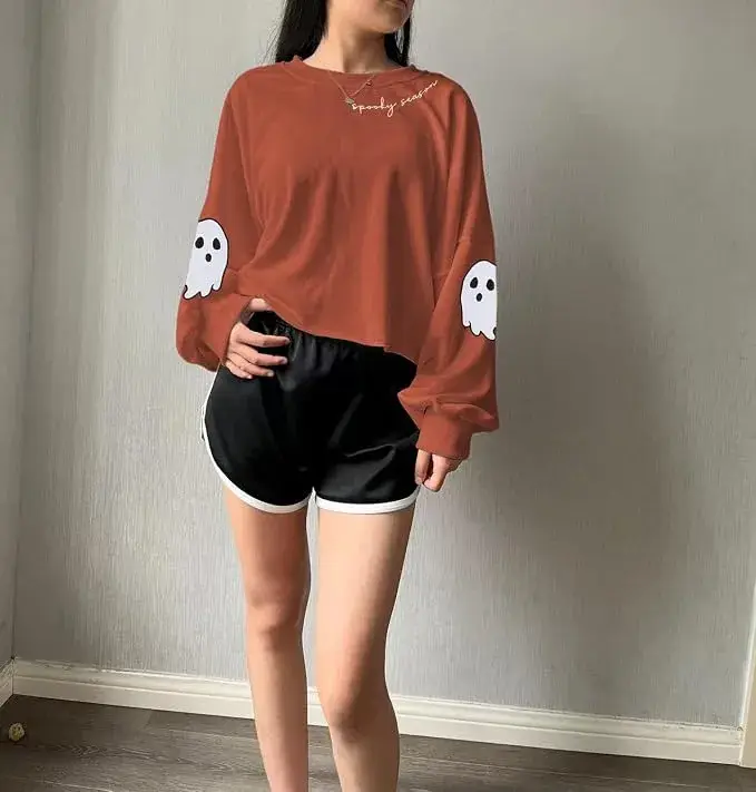 Person models a rust-red sweatshirt with white ghost patches and black athletic shorts in a gray room, creating a casual, playful look.